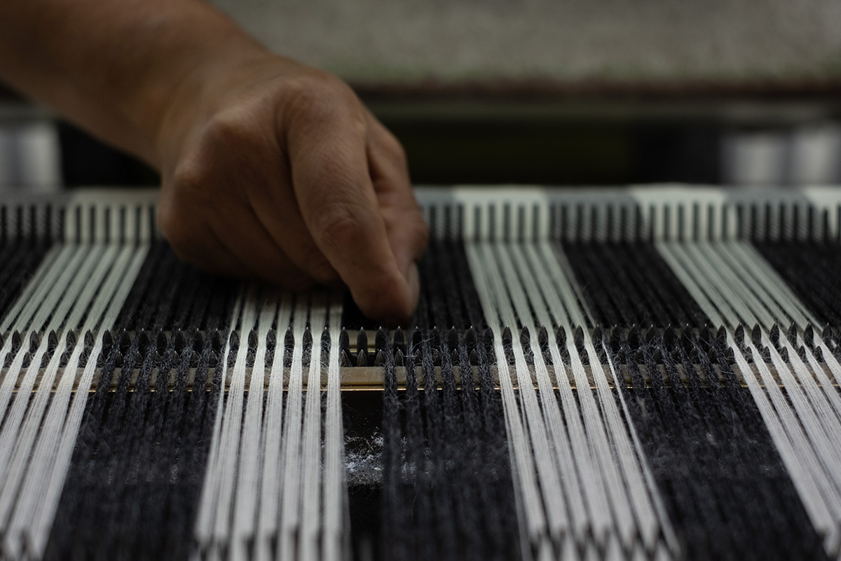 A detailed view of a hand working on a loom, with black and white threads stretched in preparation for weaving. The focus on the hand against the loom's structure conveys the precision and skill involved in textile manufacturing.