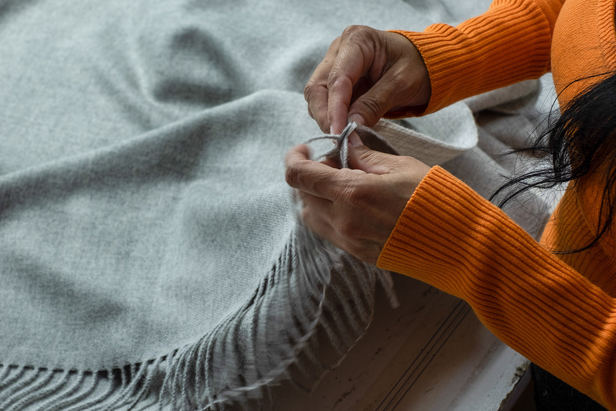 Close-up of a person's hands engaging in textile craft, with fingers intricately weaving o a gray fringed plaid. The person is wearing a vibrant orange sweater, suggesting a creative or artisanal activity in a cozy indoor setting.